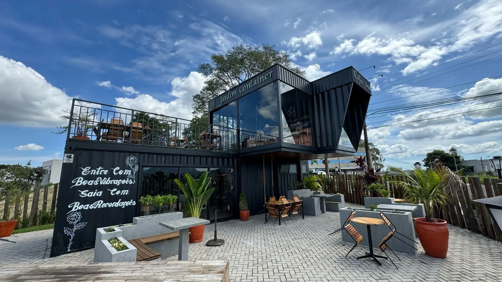 Fachada do Jardim Container em contêineres pretos empilhados, com terraço superior em guarda-corpo metálico e pátio de mesas ao ar livre