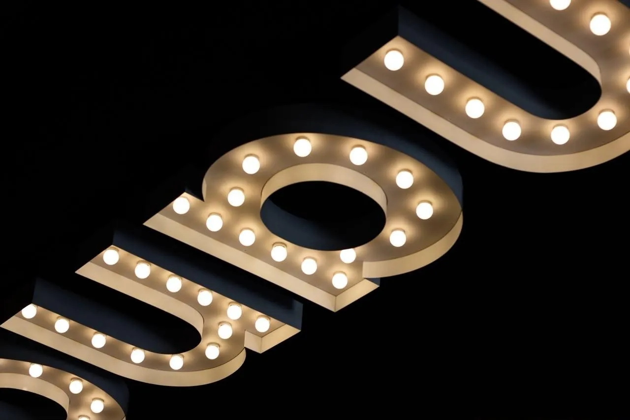 Letras de marquise em relevo dourado com lâmpadas aparentes, vistas de baixo contra o teto escuro