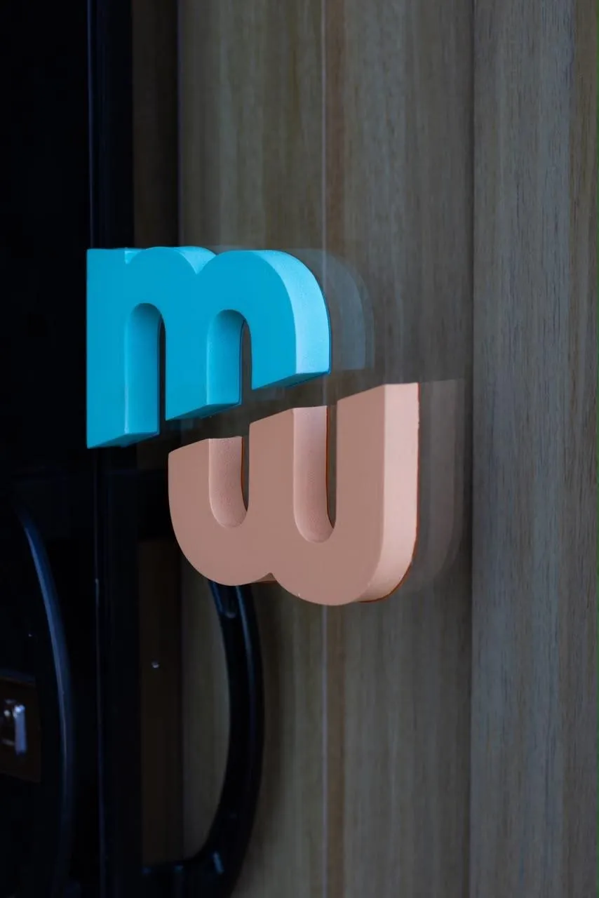 Logotipo em relevo aplicado sobre painel de madeira ripada, visto através do vidro da entrada