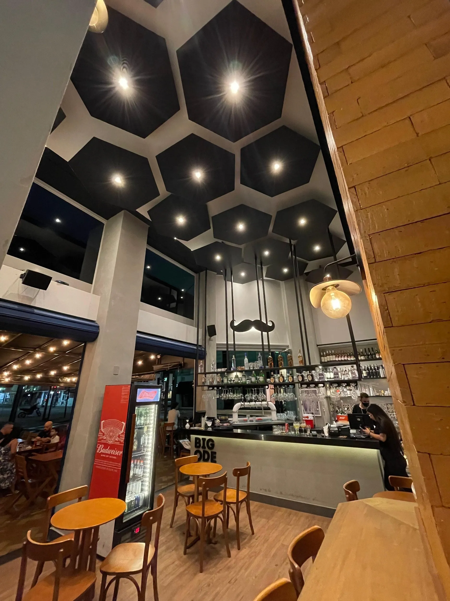 Salão com mesas altas, luminárias pendentes e o bar ao fundo
